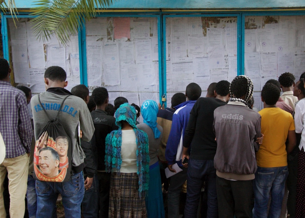 A group of people stand closely together, facing a board covered with numerous sheets of paper, appearing to examine the posted documents. Among them, someone holds a bag with a portrait on it, perhaps hinting at themes from "Modern Slavery" or referencing the "Definitive Guide.