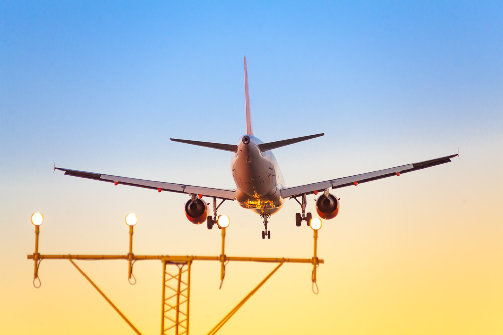 A commercial airplane with extended landing gear prepares to land against a colorful sunset sky, embodying the principles of sustainable tourism. The runway approach lights, a beacon for GSTC-certified travel, are visible in the foreground.