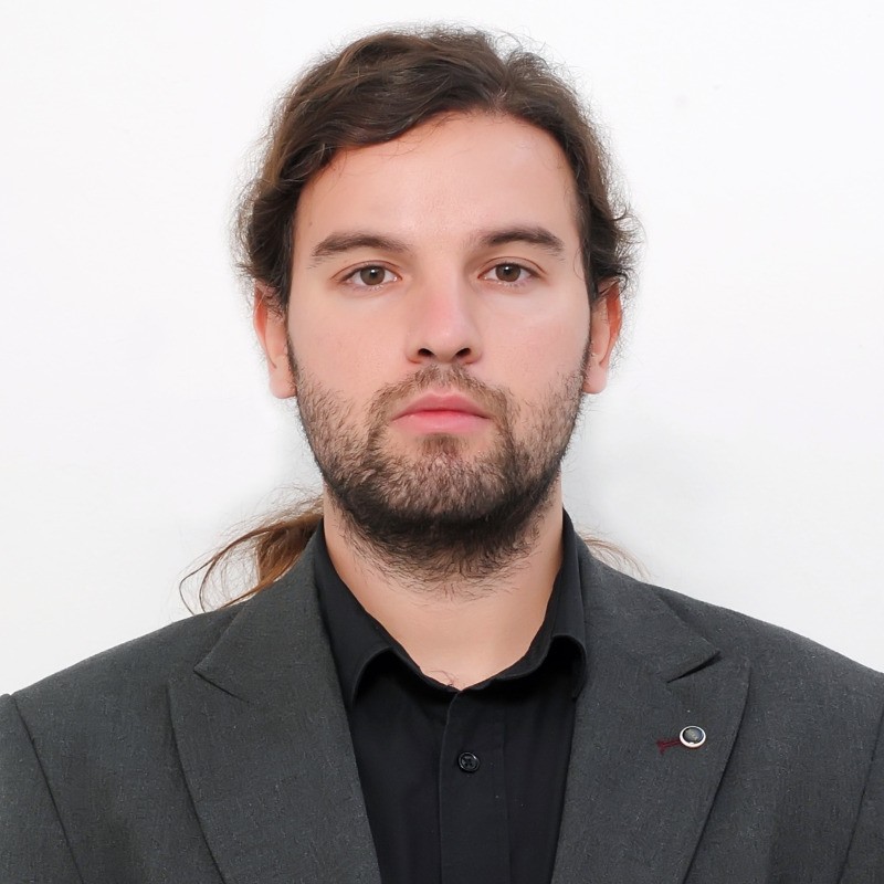 A person with long hair tied back, a beard, and a neutral expression is wearing a dark suit jacket over a black shirt. The background is plain white.