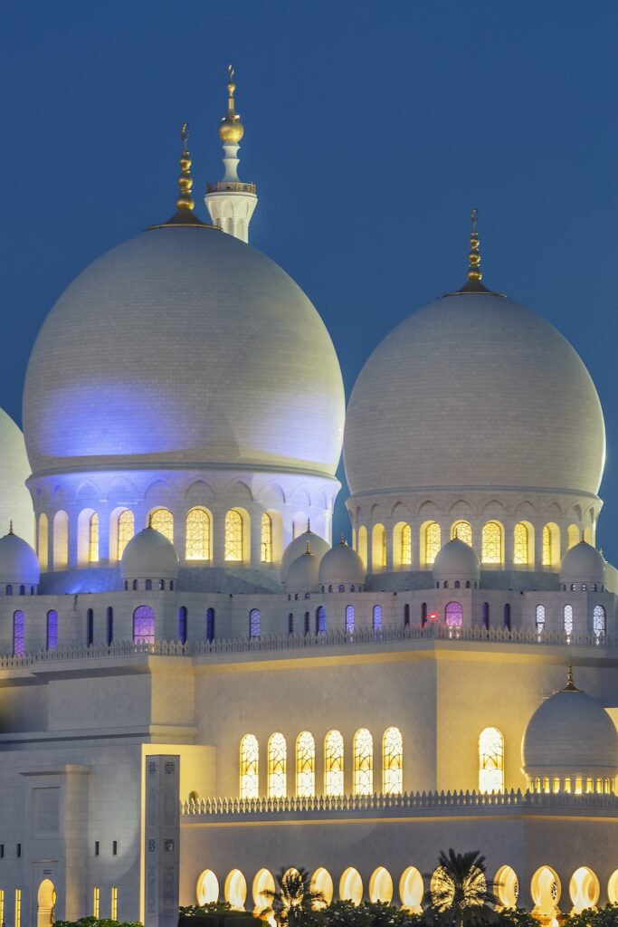 The image captures the illuminated domes of a grand mosque in the UAE at dusk. Adorned with golden finials, the domes and glowing windows embody elegance against a deep blue sky, symbolizing progress towards UAE's commitment to the Net Zero 2050 initiative. Designed by Freepik